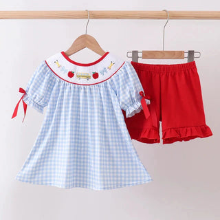 A toddler girl's two-piece clothing set hanging on wooden hangers against a plain light background. The set includes a light blue and white gingham smocked dress with red trim, a white collar with embroidered strawberries and a red apple, and red ruffle-hem shorts with red side bows.