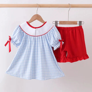 A toddler girl's two-piece outfit hanging on wooden hangers against a plain light background. The set includes a light blue and white gingham short-sleeve dress with a white Peter Pan collar trimmed in red, and matching red ruffled shorts.