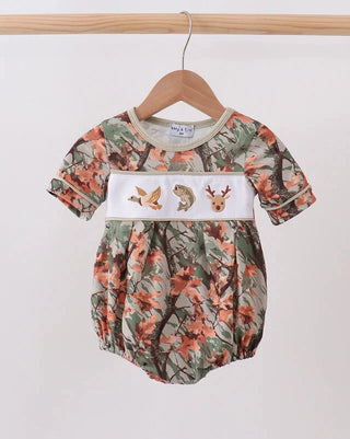 A baby short-sleeve romper with a woodland camouflage print, featuring a white smocked chest panel decorated with embroidered deer and bird motifs, hanging on a wooden hanger against a plain light background.