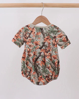 A short-sleeve baby romper with an all-over branch and leaf print in green, brown, and orange, hanging on a wooden hanger against a plain white wall.
