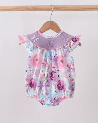 A baby romper with a purple floral print and a lavender Peter Pan collar, hanging on a wooden hanger from a wooden clothes rack against a plain white wall.