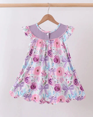 A toddler girl's floral print dress hanging on a wooden hanger against a plain light background. The dress features a lavender collar, ruffled cap sleeves, and a watercolor-style print of pink and purple roses with bows on a white base.