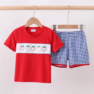 A children's two-piece clothing set hanging on a wooden rod. The set includes a bright red short-sleeve t-shirt with a white horizontal chest band featuring embroidered red and green apples, and matching blue-and-white gingham shorts with red trim.