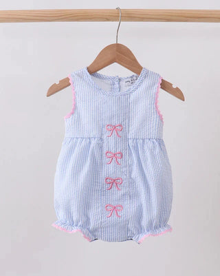 A sleeveless baby romper in light blue and white gingham fabric, featuring pink embroidered bows down the front and pink piped trim along the armholes and ruffled hem, hanging on a wooden hanger.