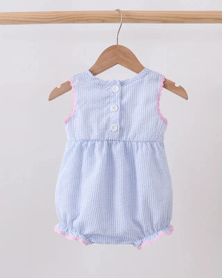 A sleeveless baby romper in a light blue and white gingham pattern, featuring a button-down front and pink ruffle trim, hanging on a wooden hanger from a wooden clothes rack against a plain light background.