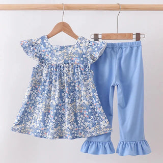 A toddler girl's two-piece clothing set hanging on wooden hangers against a plain white wall. The set includes a light blue floral print smocked dress with ruffled sleeves and matching light blue ruffled capri pants.