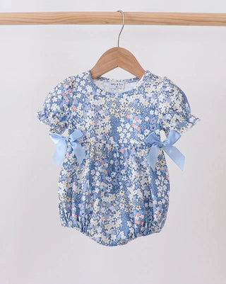 A baby girl's short-sleeve floral romper in light blue, white, and soft pink, hanging on a wooden hanger from a wooden clothes rack. The romper features ruffled sleeves with light blue ribbon bows on the sides.
