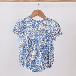 A baby girl's short-sleeve romper in a light blue and white floral print, hanging on a wooden hanger from a wooden clothes rack against a plain light gray wall.