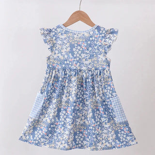 A blue and white floral print baby dress with ruffled sleeves and gingham side pockets, hanging on a wooden hanger against a plain light gray wall.