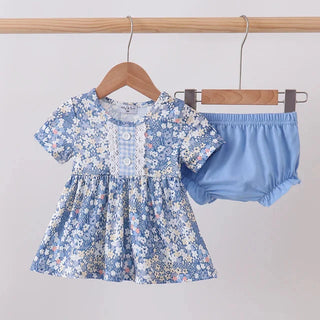 A baby girl's two-piece outfit hanging on wooden hangers from a clothing rack. The set includes a short-sleeve smocked dress in a light blue and white floral print with a lace yoke, and matching light blue bloomers.