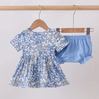 A toddler girl's two-piece outfit hanging on wooden hangers against a plain light gray wall. The set includes a short-sleeve dress with a blue, white, and yellow floral print, and a pair of solid light blue bloomers.
