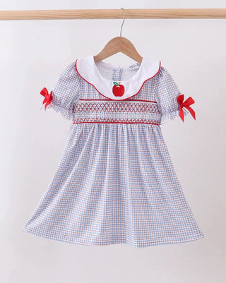 A toddler girl's short-sleeve dress with a white and blue gingham pattern, featuring red smocked detailing, a white collar with red trim, red bow accents on the sleeves, and a red apple applique at the neckline, hanging on a wooden hanger against a plain light background.