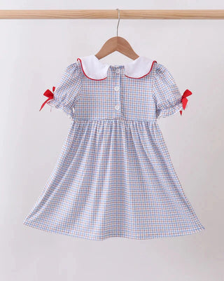 A toddler girl's short-sleeve dress with a blue, white, and red gingham pattern, featuring a white Peter Pan collar with red trim and red ribbon bows on the sleeves, hanging on a wooden hanger.