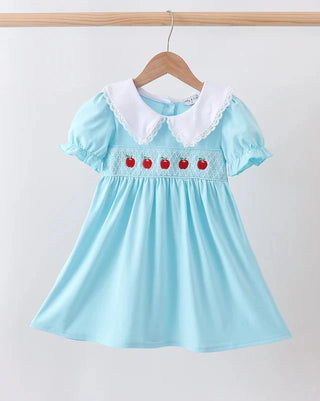 A light blue girl's dress with a white Peter Pan collar and embroidered red apple details, hanging on a wooden hanger against a plain white wall.
