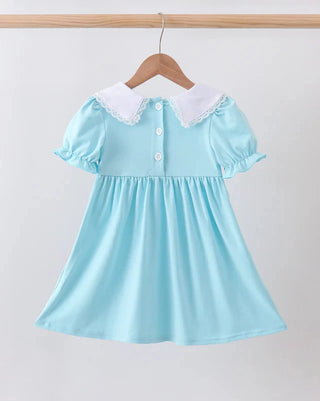 A light blue girl's dress with a white lace Peter Pan collar and short puff sleeves, hanging on a wooden hanger against a plain light gray background.
