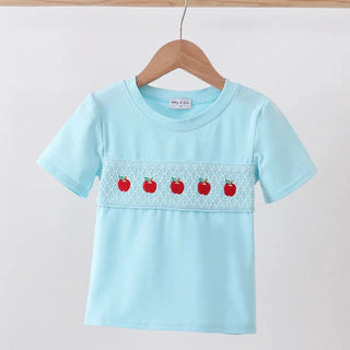 A light blue short-sleeve boy t-shirt hanging on a wooden hanger. The shirt features a decorative horizontal band across the chest with white embroidered geometric patterns and five red embroidered apples with green stems.