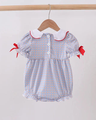 A baby girl's short-sleeve romper with a blue, white, and red gingham check pattern, featuring a white Peter Pan collar with red trim, buttoned front, and red ribbon bows on the sleeves, hanging on a wooden hanger.