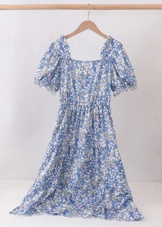A blue and white floral print dress with puff sleeves and a square neckline, hanging on a wooden hanger against a plain white wall.