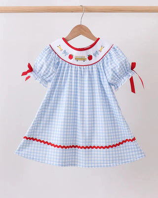 A light blue and white gingham dress hanging on a wooden hanger against a plain light background. The dress features a white collar with red trim, embroidered details, and red ribbon bows on the sleeves.
