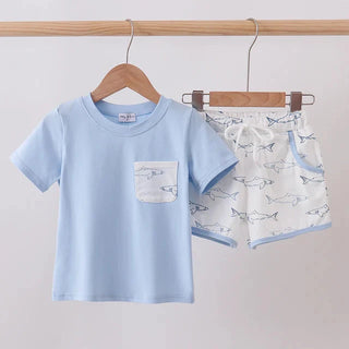 A children's two-piece outfit hanging on wooden hangers from a wooden rod. The set includes a light blue short-sleeve t-shirt with a white chest pocket and matching white shorts printed with small blue fish, both featuring light blue trim.