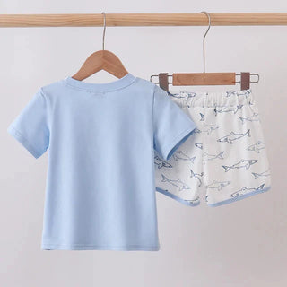 A two-piece children's outfit hanging on wooden hangers from a wooden rod. The set includes a light blue short-sleeve t-shirt and white shorts with a blue shark pattern.