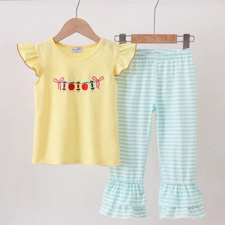 A children's two-piece outfit hanging on wooden hangers against a plain light background. The set includes a pale yellow short-sleeve top with ruffled sleeves and a colorful embroidered holiday garland graphic across the chest, paired with mint green and white striped ruffled capri pants.