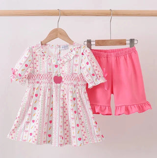 A toddler girl's two-piece clothing set hanging on wooden hangers against a plain light background. The set includes a short-sleeve smocked dress in white with pink and green floral/heart patterns, and a pair of bright pink ruffle-trimmed shorts.