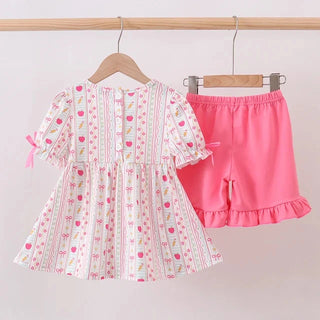 A toddler girl's two-piece clothing set hanging on wooden hangers against a plain light background. The set includes a short-sleeve dress with a white base, pink and green vertical stripes, and a small pink strawberry print, paired with bright pink ruffled shorts.