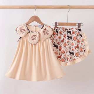 A toddler girl's two-piece outfit hanging on wooden hangers from a light wood rod. The set includes a cream-colored sleeveless dress with ruffled sleeves featuring embroidered horse motifs, and matching ruffled shorts with a multicolor print of horses and flowers on a cream background.