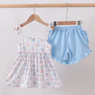 A toddler girl's two-piece outfit hanging on wooden hangers from a wooden rod. The set includes a sleeveless white dress with a pink and blue floral print and ruffle trim, and light blue shorts with ruffle trim.