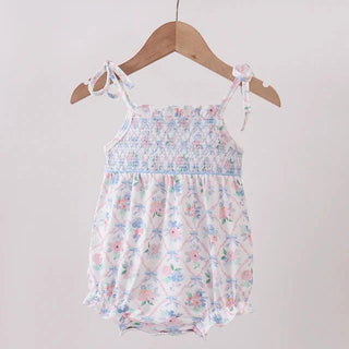 A baby girl's smocked romper with a delicate pink, blue, and green floral print, hanging on a wooden hanger from a light wood clothing rack against a plain off-white wall.