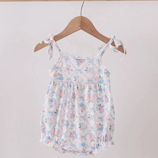 A toddler girl's sleeveless romper with a delicate pastel floral print, hanging on a wooden hanger from a light wood clothing rack against a plain off-white wall.