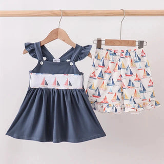 A toddler girl's two-piece outfit hanging on wooden hangers against a light wall. The set includes a slate blue sleeveless dress with flutter sleeves and a white band featuring a sailboat print, paired with a matching white ruffled skirt covered in a colorful sailboat pattern.