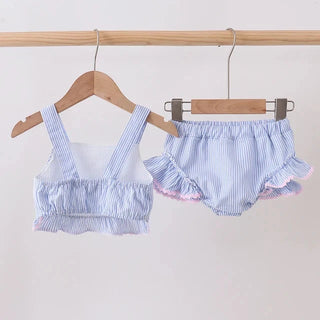 A two-piece baby swim set consisting of a light blue and white striped ruffled crop top and matching ruffled bloomers, hanging on wooden hangers from a wooden clothing rack against a plain off-white wall.