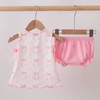 A baby girl's two-piece outfit hanging on wooden hangers from a wooden rod. The set includes a white sleeveless top with pink bow patterns and ruffle trim, and matching pink bloomers.