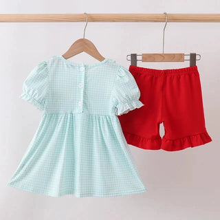 A toddler girl's two-piece clothing set hanging on wooden hangers from a wooden rod. The set includes a light mint green and white gingham short-sleeve top with ruffled sleeves and buttons on the back, and solid red ruffled shorts.
