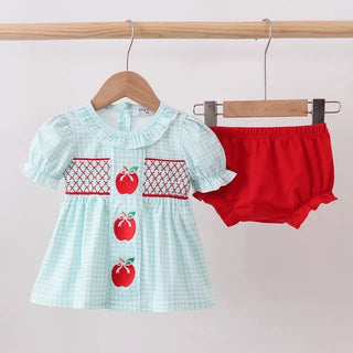 A baby girl's two-piece outfit hanging on a wooden hanger from a clothing rack, featuring a light blue and white gingham dress with red apple motifs and a red smocked chest panel, paired with bright red bloomers.