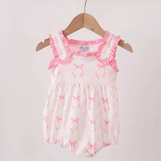 A baby romper in white with pink bow prints and pink ruffle trim, hanging on a wooden hanger from a light wood rod against a plain off-white wall.