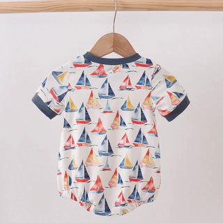 A baby short-sleeve romper hanging on a wooden hanger from a wooden clothes rack. The romper has an all-over sailboat print on a white background with navy blue trim on the collar and cuffs.