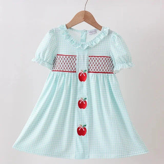A light mint green and white gingham baby dress hanging on a wooden hanger against a plain light beige wall. The dress features short puff sleeves, a ruffled collar, red and white embroidered chest panels, and three red apple appliqués down the front.