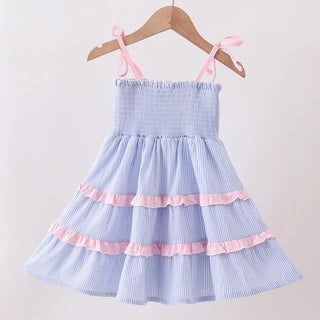 A light blue and white gingham tiered baby dress with pink ruffle accents and tie straps, hanging on a wooden hanger against a plain white wall.