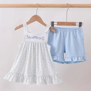 A toddler girl's two-piece summer outfit hanging on wooden hangers against a plain light background. The set includes a white sleeveless smocked dress with tiny blue floral prints and 'Sweet Princess' embroidered on the chest, paired with light blue ruffled shorts.