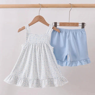 A toddler girl's two-piece clothing set hanging on wooden hangers from a light wood rod. The set includes a white sleeveless smocked dress with a tiny blue floral pattern and ruffled hem, paired with light blue ruffled shorts.
