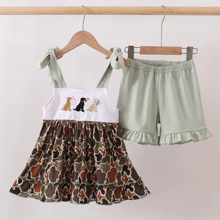 A toddler girl's two-piece outfit set hanging on wooden hangers against a plain light background. The set includes a sleeveless ruffled top with a white yoke featuring dog illustrations and a brown camouflage-patterned skirt, paired with light sage green ruffled shorts.