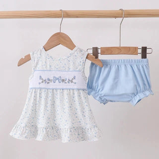 A baby girl's two-piece outfit set hanging on wooden hangers from a wooden rod. The set includes a white sleeveless dress with a tiny blue floral print, a light blue waistband with embroidered blue bows, and a pair of light blue bloomers.