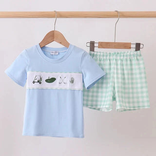 A children's two-piece clothing set hanging on wooden hangers against a plain light background. The set includes a light blue short-sleeve T-shirt with a white chest panel featuring golf-themed illustrations, and light green and white gingham shorts.