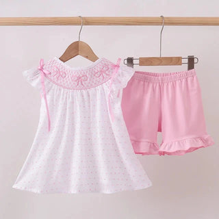 A two-piece toddler girl's outfit set hanging on wooden hangers against a light pink wall. The set includes a white short-sleeve dress with tiny pink polka dots, a pink embroidered collar with bow details, and matching pink ruffle-trimmed shorts.