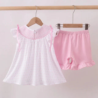 A two-piece baby girl's outfit set hanging on wooden hangers from a wooden clothes rack. The set includes a white short-sleeve dress with tiny pink polka dots and a pink patterned collar with tie details, and a pair of pink shorts with a ruffled hem.