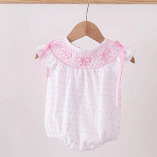 A white baby romper with pink polka dots, featuring a pink embroidered bow pattern on the yoke and matching pink ribbon ties on the shoulders, hanging on a wooden hanger.