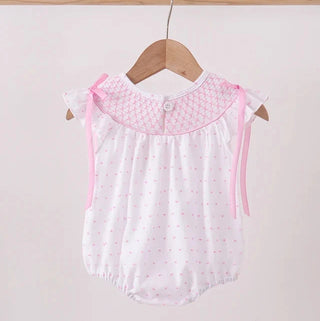 A baby girl's sleeveless romper in white with a pink polka dot pattern, featuring a textured pink collar and pink ribbon ties, hanging on a wooden hanger.
